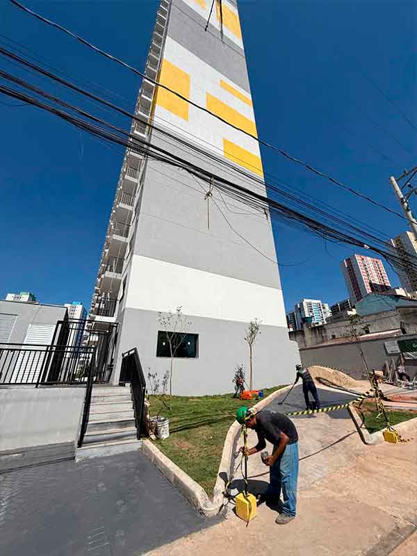 Fachada lateral de prédio alto com pintura em tons de cinza, branco e amarelo, com operários trabalhando na base.